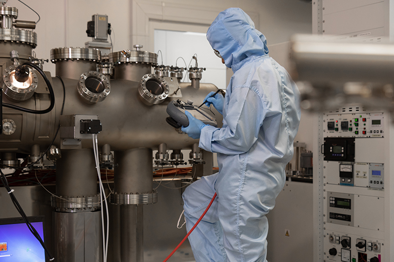 person in a light blue lab suit with their back to us standing at a grey item in a laboratory