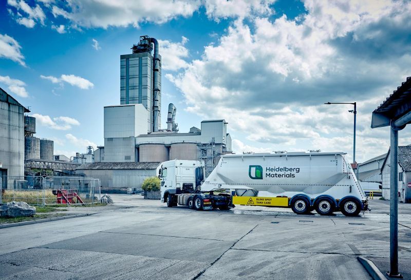 Cement tanker on a concrete parking area with industrial buildings behind under a blue sky with clouds