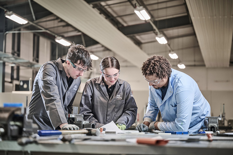 Two apprentices with a teacher in overcoats, protective glasses and gloves in a large workshop area, leaning over some plans
