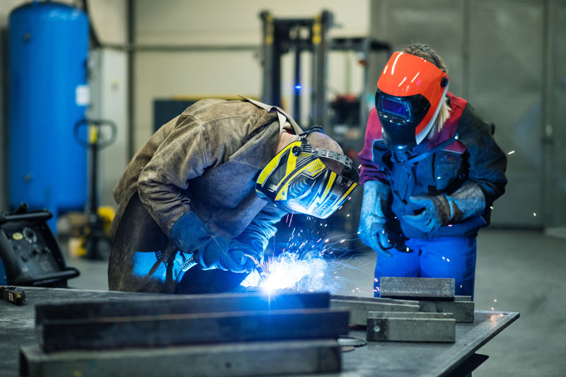 Blonde woman works as a welder in workshop, operating welding machine, wearing protective clothing and a welding mask
