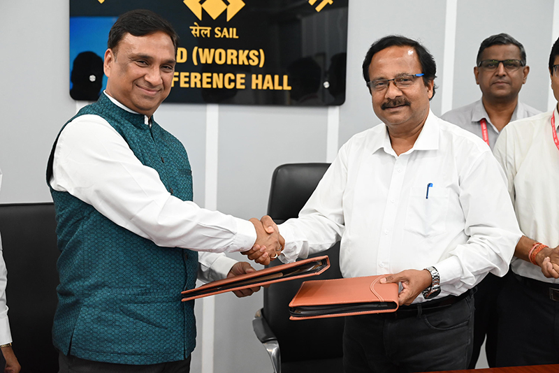 two smiling people exchange brown document wallets and shake hands while others watch on. There is a sign on the wall behind indicating this is housed in a company conference hall