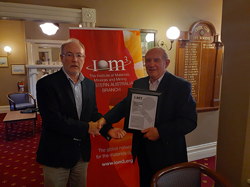 two men in dark jackets stand in front of an IOM3 po-up banner, man to the right holds a certificate