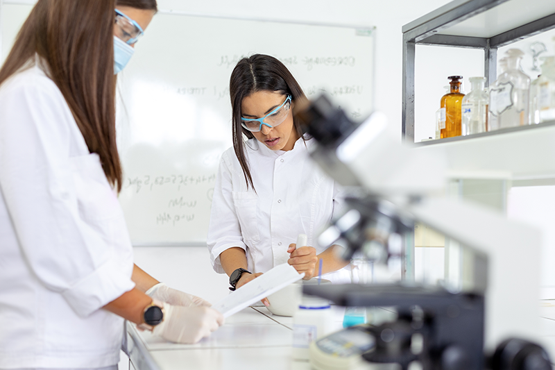 Stock image of two female researchers working in a lab space with a microscope and mortar and pestle