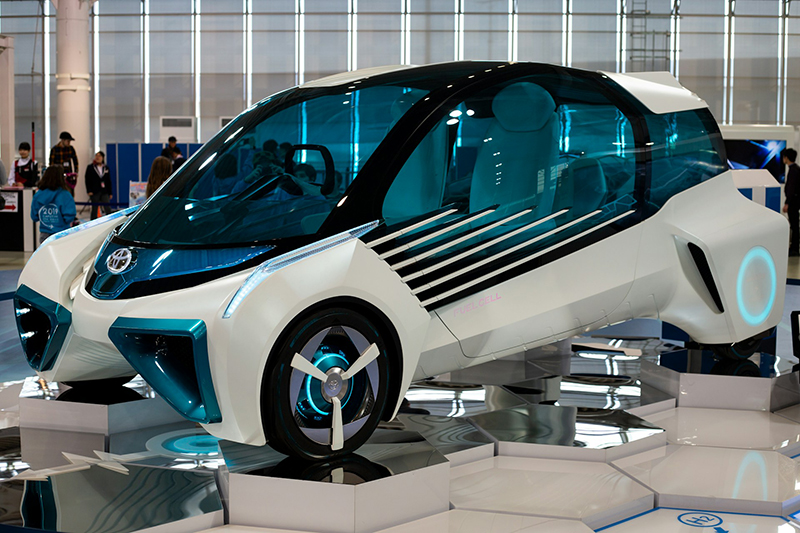 A Toyota hydrogen fuel cell concept vehicle on display, the vehicle has a white body with blue around the headlights, it is held at an angle with the front downwards, people ar visible behind