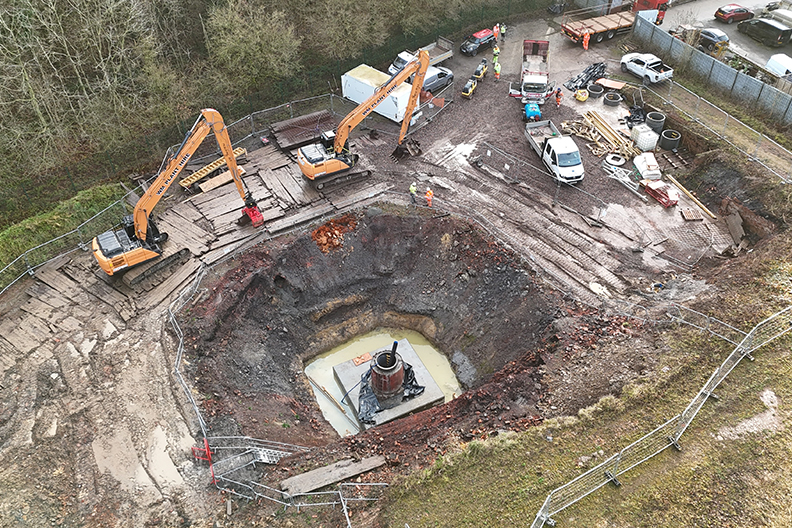 Picture of a large pit about 10-20m deep from a birds eye view which has a concrete square at the bottom of the pit, with construction vehicles around the sides
