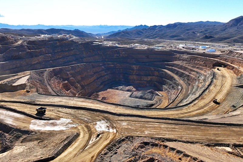 A view of a huge open pit mine on a sunny day with giant dumper trucks that look tiny and dirt circular tracks that form concentric circles around the outside