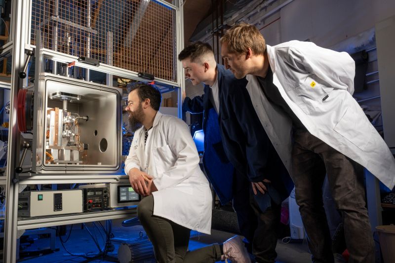 three spope in lab coats looking intently at a glowing glass and metal hood. one is seated the others stand to the right