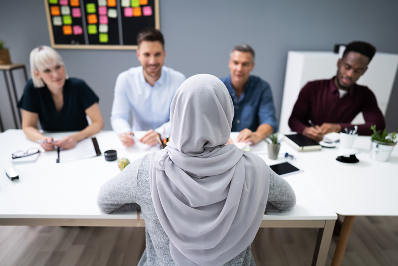 single person in headscarf faces a panel of four other people at a table which has notebooks and coffee cups