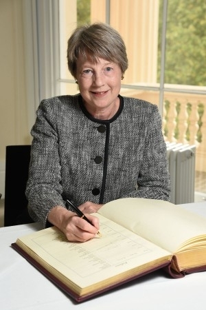 Rachel Williams signing the RAEng Fellows book, she is seated behind a desk and looks to the camera with a smile, pen poised over the book, she wears a dark jacket
