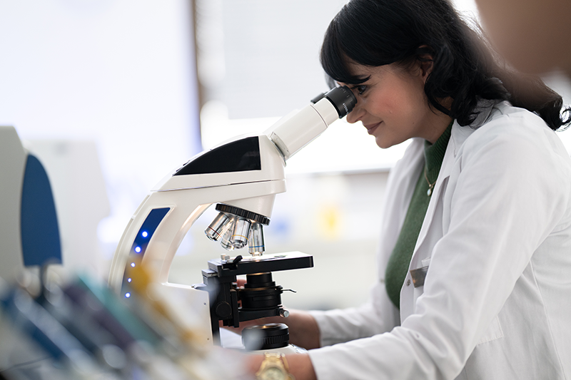 Stock image of a female researcher in a lab coat and goggles, looking down a microscope