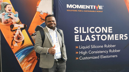 Chris Nwosu in a checked jacket and white shirt standing in front of a Momentive banner showing a car