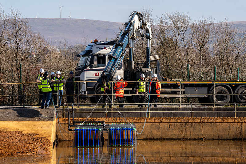 two groups of people by a white lorry and a crane lover blue items into water