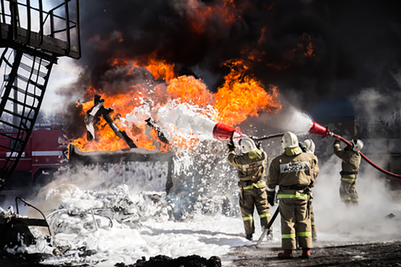 Fire fighters holding two large hoses to put out a fire in an industrial setting, one clearly with a lot of foam