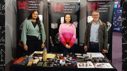 Three people standing behind a desk with materials. They are at the GUTS event in Sheffield and have SMEA pop up banners behind them.