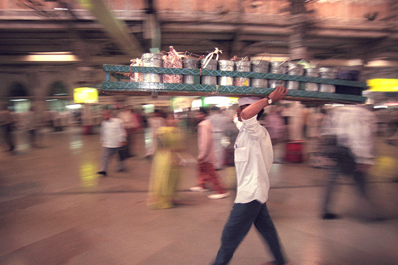 man walking with a large metal tray balanced on his head containing tins, focus is on him, there are blurred people behind
