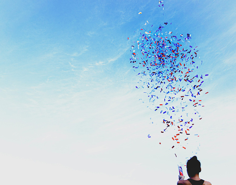 A person, seen from the rear fires a confetti cannon into a blue and white sky