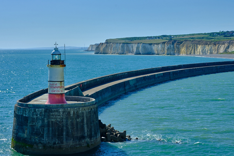 The entrance to Newhaven harbor, East Sussex, UK, and its breakwater and lighthouse. Clear blue sky and water with white cliffs in the background.