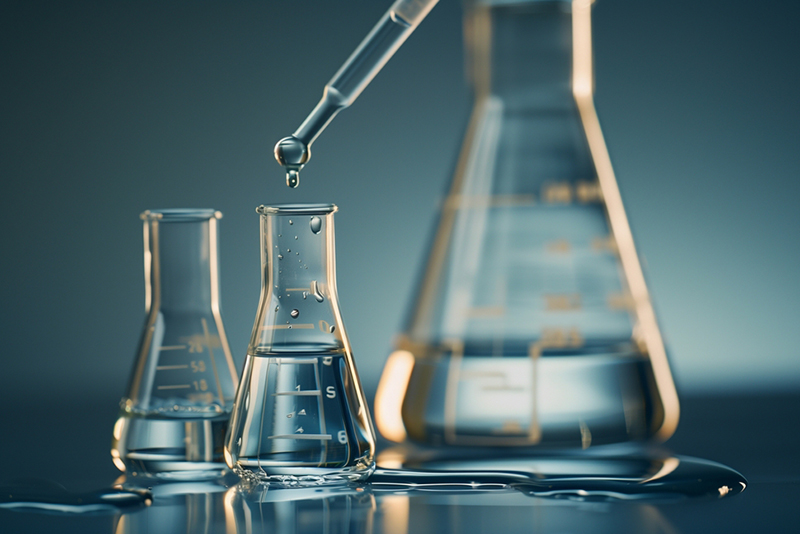 Large flas with distillation tube, clear liquid drips from the tube into a smaller flask, Another flask sits to the right of the small coecting one and there is liquid spilt around the base of both