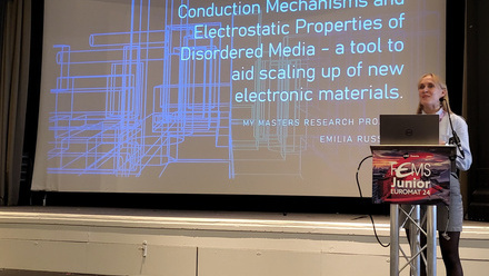 Large screen with presentation slide to the leaft and female speaker to the right in a light coloured shirt and grey jeans with trainers