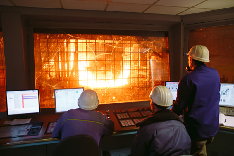 Workers in hard hats and jackets sit in a control room with monitors, watching a giant industrial furnace through the window