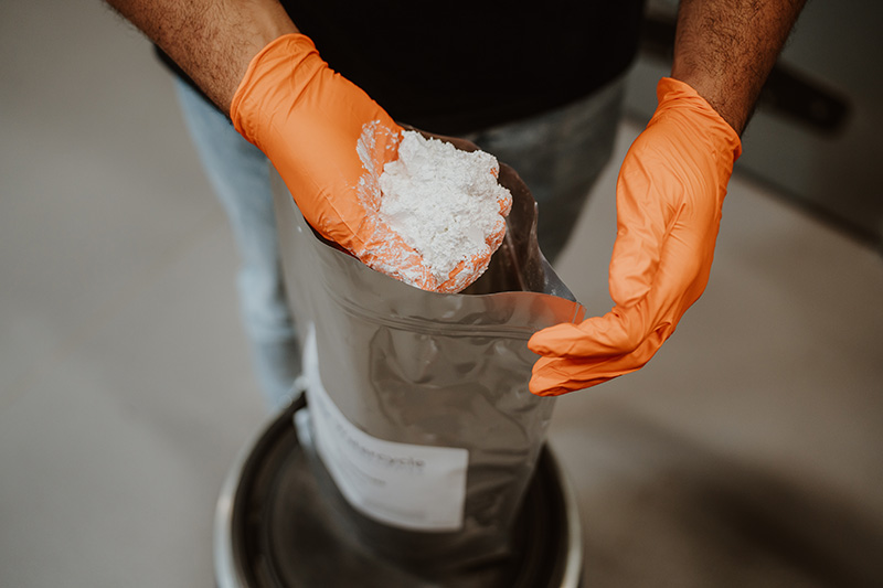 person wearing orange gloves holds white powder above a foil packet in a bucket