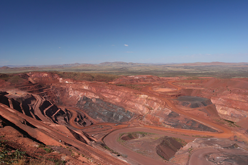 A view from the top of an open pit mine in Western Australia, with dirt roads snaking down into the pit and hills in the distance