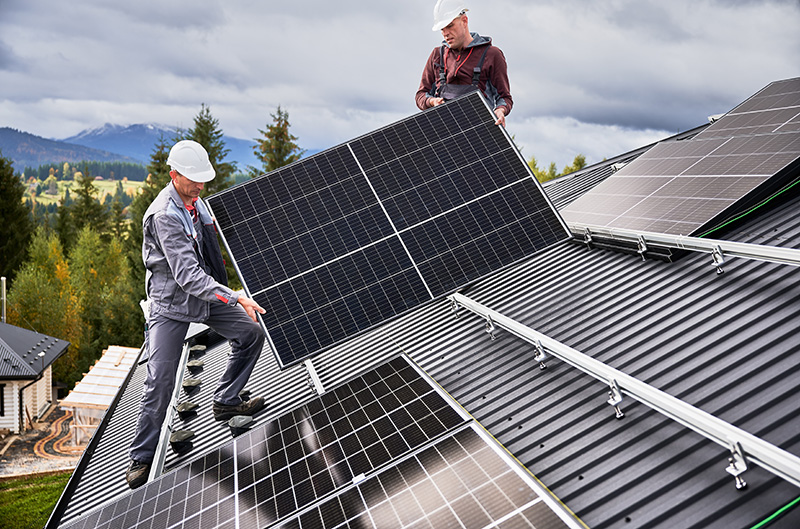 two men in hard hats install solar panels on a sloping roof, trees are visible in the distance