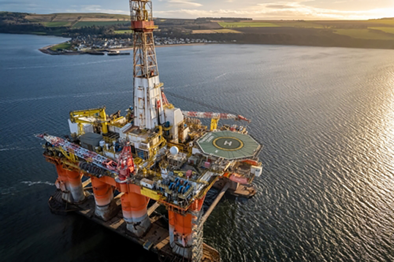 Offshore oil platform seen from above near the coast with green fields in the background on a sunny day