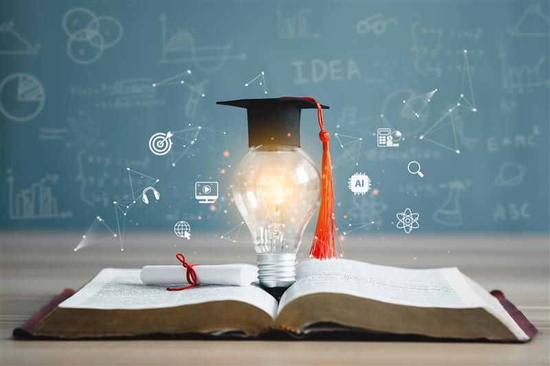 blackboard to rear with chalked symbols, on desk at front an open book, a graduation scroll and a lit lightbulb with morterboard and orange tassle