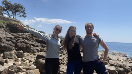 Three people with linked arms standing on a rocky area with sea behond and a slope to the right with one tree