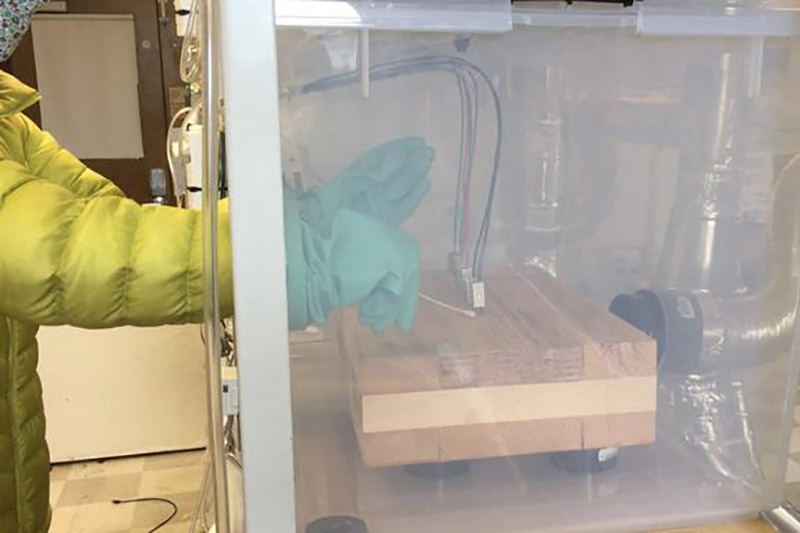 researcher in a yellow suit reaches into a hood to work on a wooden block