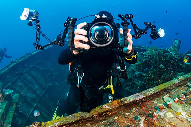 A diver holding an underwater camera while swimming just above a metal shipwreck