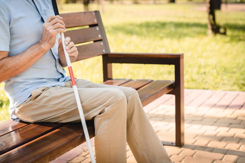 visually impaired blind senior man holding white and red guide cane walking stick, sitting outdoors on a wooden bench in park