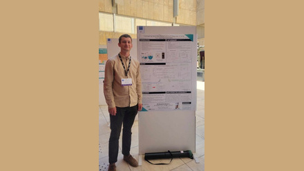 A male in a beige shirt and dark trousers with brown shoes to the left of a poster presentation board