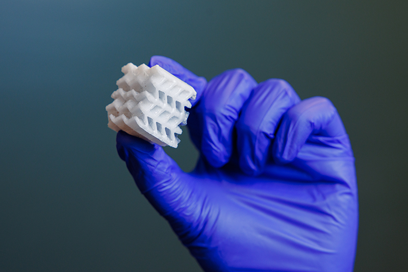 A gloved hand holding up and squeezing a geometric latticed 3D printed block that has alternating filled squares and holed squares 