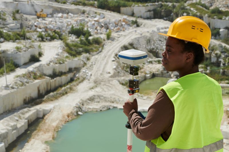 female in high-vis and hard hat holds a surveying tool at the top of an open cast mine