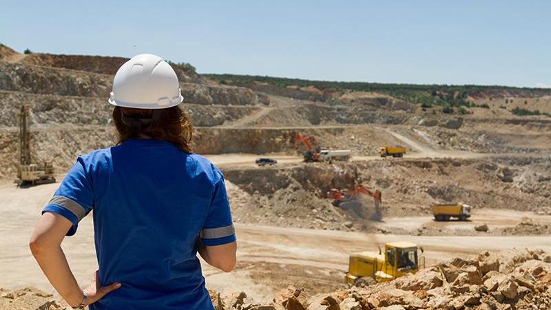 female in hard hat with blue uniform looks over an open cast mine with trucks