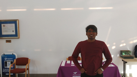Aston University logo on a white wall with a red cloth covered table and a person standing in front in a red top with black jeans and white trainers