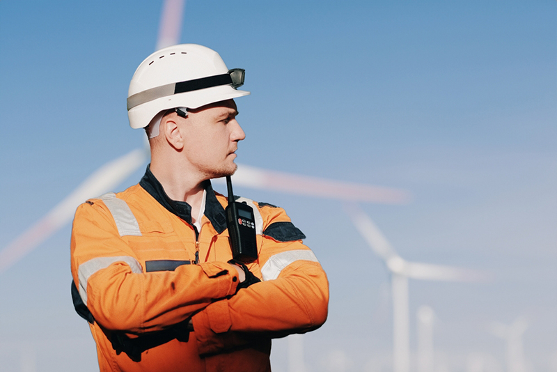 A Man In A Working Overall Boiler Suit With A Radio And Safety Helmet With A Blurred Wing Generators In The Background