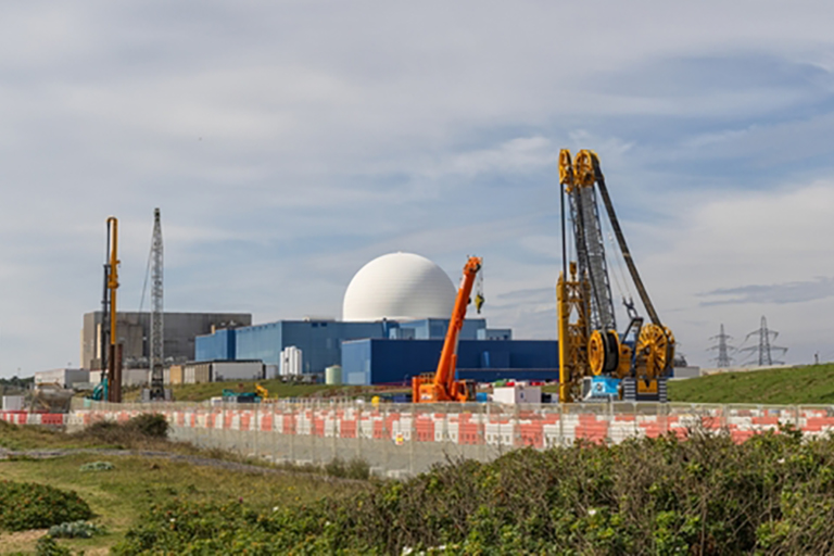 Some large construction equipment behind a fenceline with buildings in the middle-background, one with a semi-circular large globe on top, and pylons in the distance