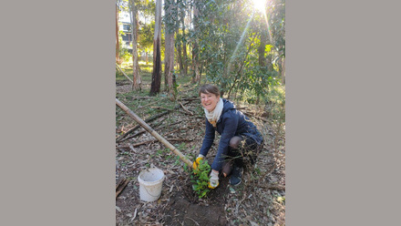 Female in a forest with a stick and bucket, kneeling on the ground smiling at the camera