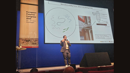 Central large screen showing a presentation, a speaker stands in front of the screen in trousers, glasses and jacket