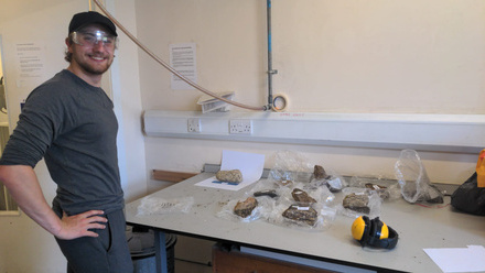 man in dark clothing with goggles next to a table displaying geological samples