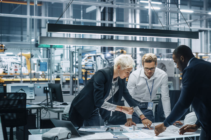 three people collaborating while standing at a table with a manufacturing set up behind, a robot arm is visible