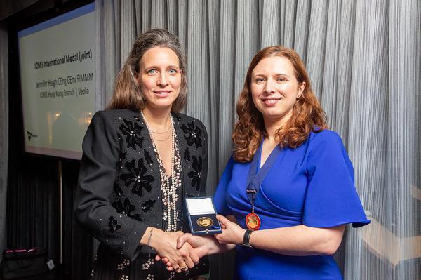 Two females shaking hands while exchanging a boxed medal, female on the left in black, and right in blue